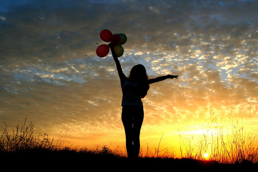 Mujer a contraluz de un atardecer con globos en la mano y elevándolos al cielo. 