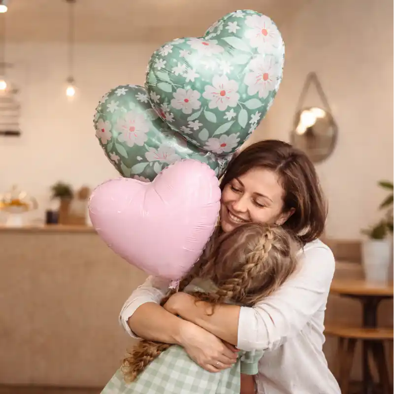 Madre e hija abrazándose con globos de corazón pastel y globos florales, decoración elegante para el Día de la Madre en interior acogedor