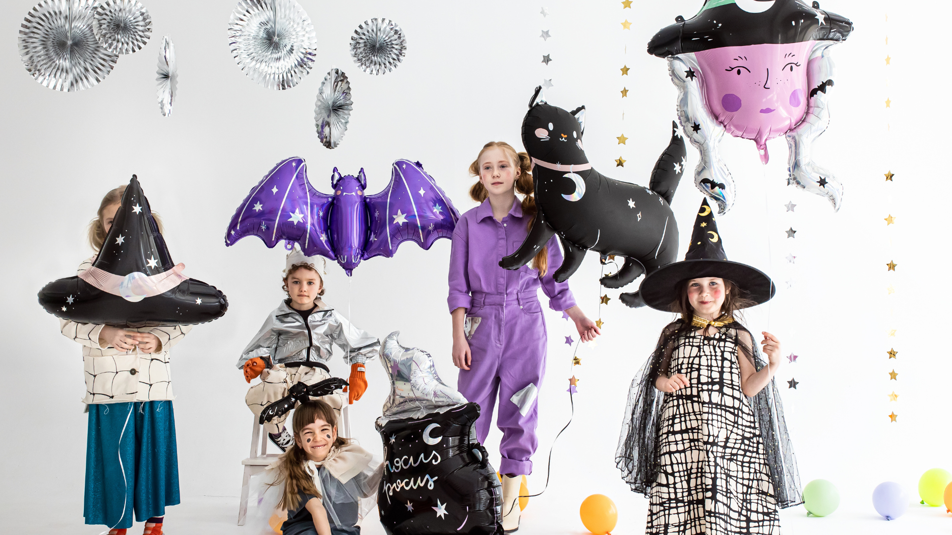 La imagen muestra a un grupo de niños disfrazados con temática de Halloween, rodeados de globos y decoraciones festivas. Algunos llevan disfraces de brujas y criaturas nocturnas, como un murciélago y un gato negro. Hay globos con formas de sombreros de bruja, un caldero y estrellas doradas colgando, creando un ambiente festivo y mágico.